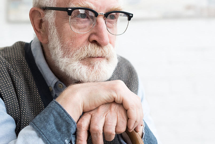 Sitting older man with a worried expression, holding a cane with both hands and resting his chin on his hands