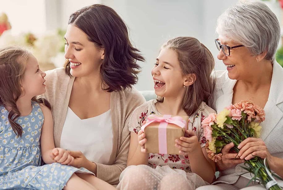 Grandmother, mother, and two daughters smiling and celebrating with gifts and flowers.