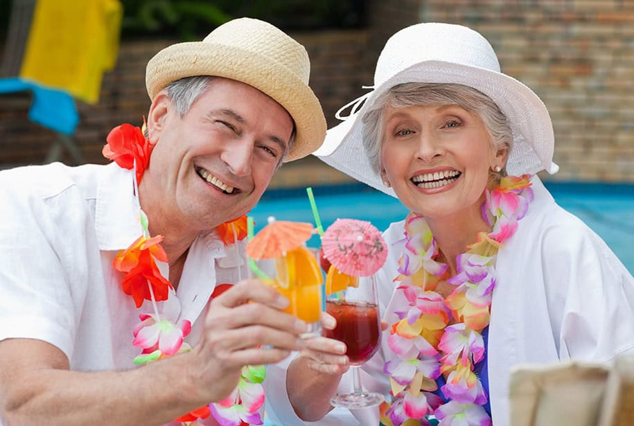 Smiling older couple sitting by a pool wearing hats and flower necklaces while holding drinks with little umbrellas in them