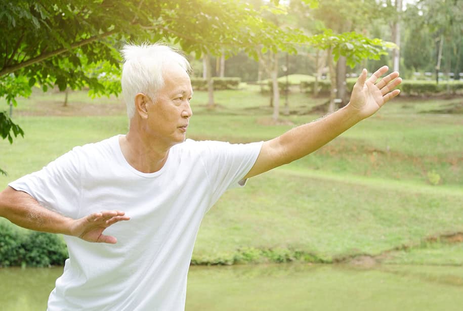 Older man in loose white clothing doing tai chi in a park, leaning slightly forward with one arm extended