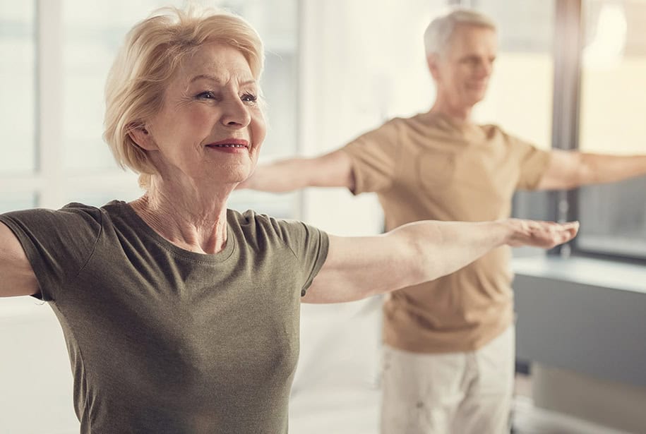 Man and woman standing with their arms stretched out to their sides in a room lined with windows