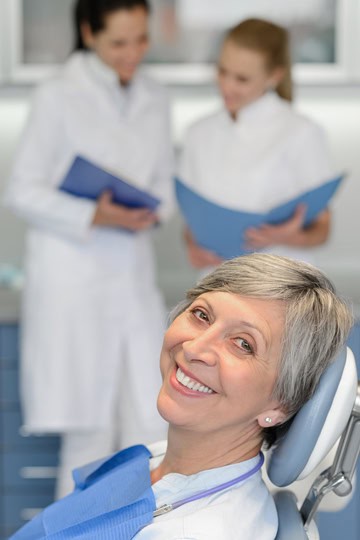 Smiling senior woman sitting in a dental chair while two dentists review her treatment in the background.