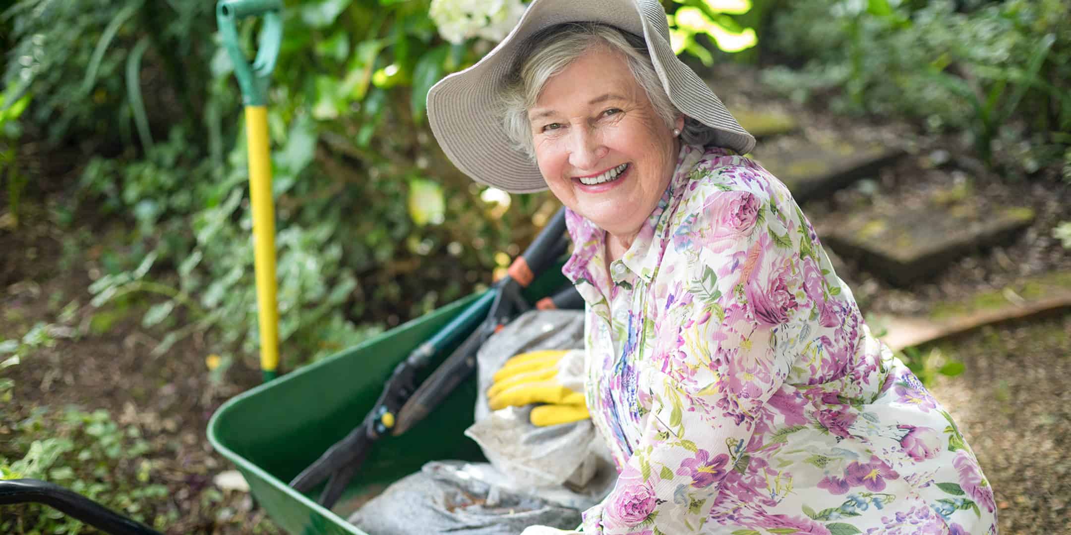 Smiling senior woman wearing a sun hat and floral shirt, gardening with a wheelbarrow and tools outdoors.