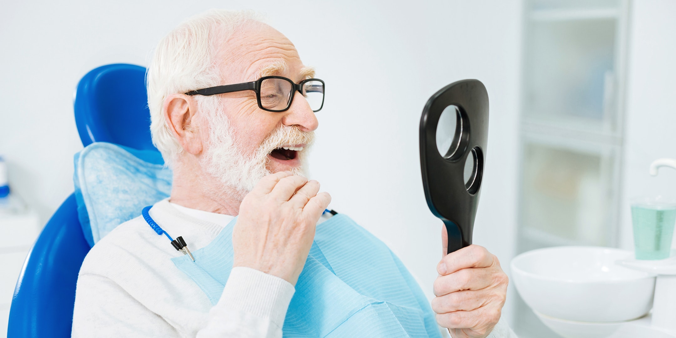 Senior man in a dental chair happily examining his teeth in a handheld mirror.