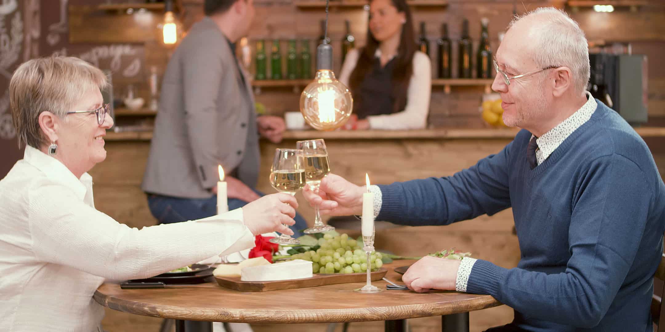 Senior couple toasting with wine at a cozy restaurant with candlelight and a cheese platter.