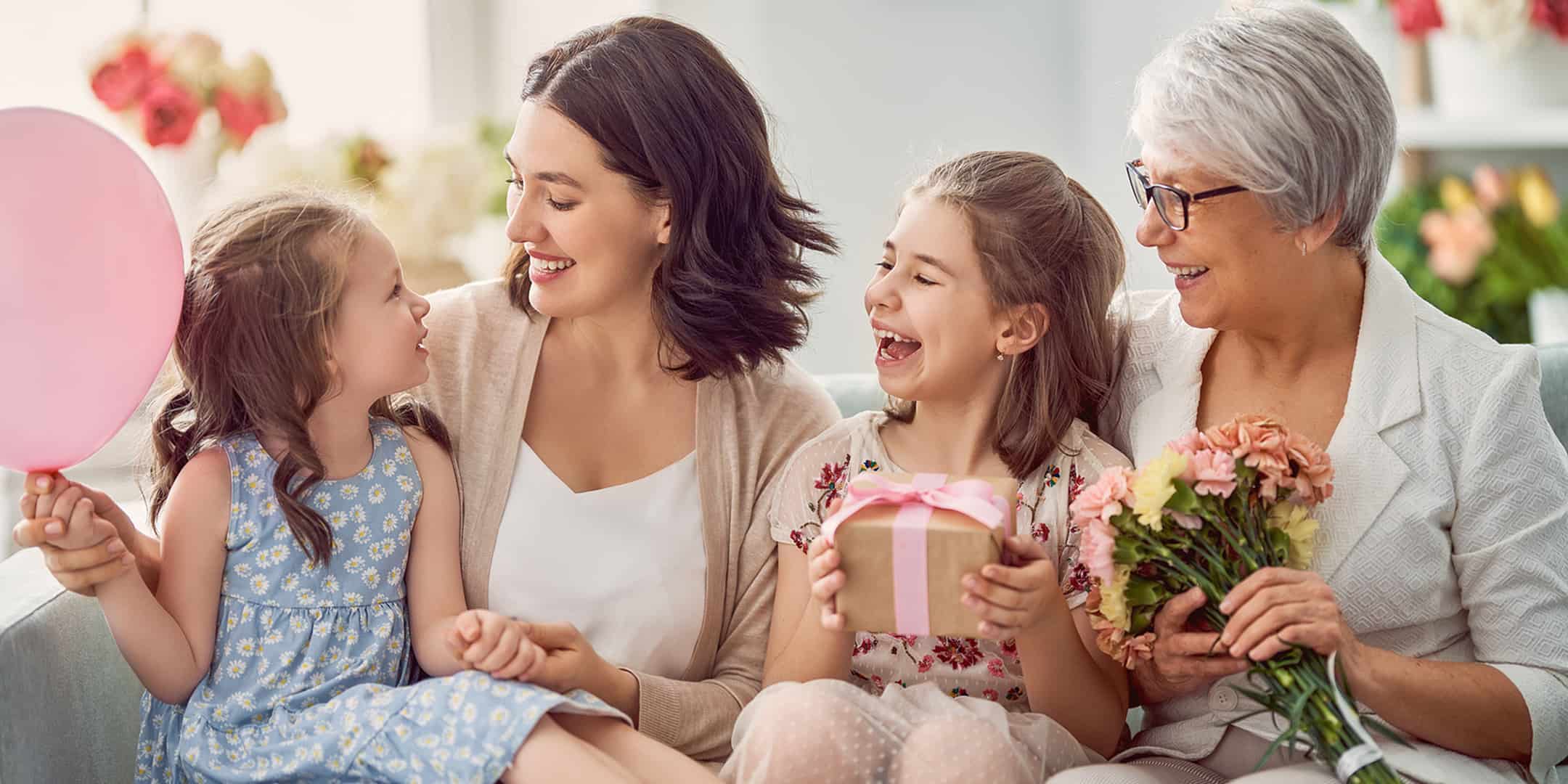 Grandmother, mother, and two daughters smiling and celebrating with gifts and flowers.