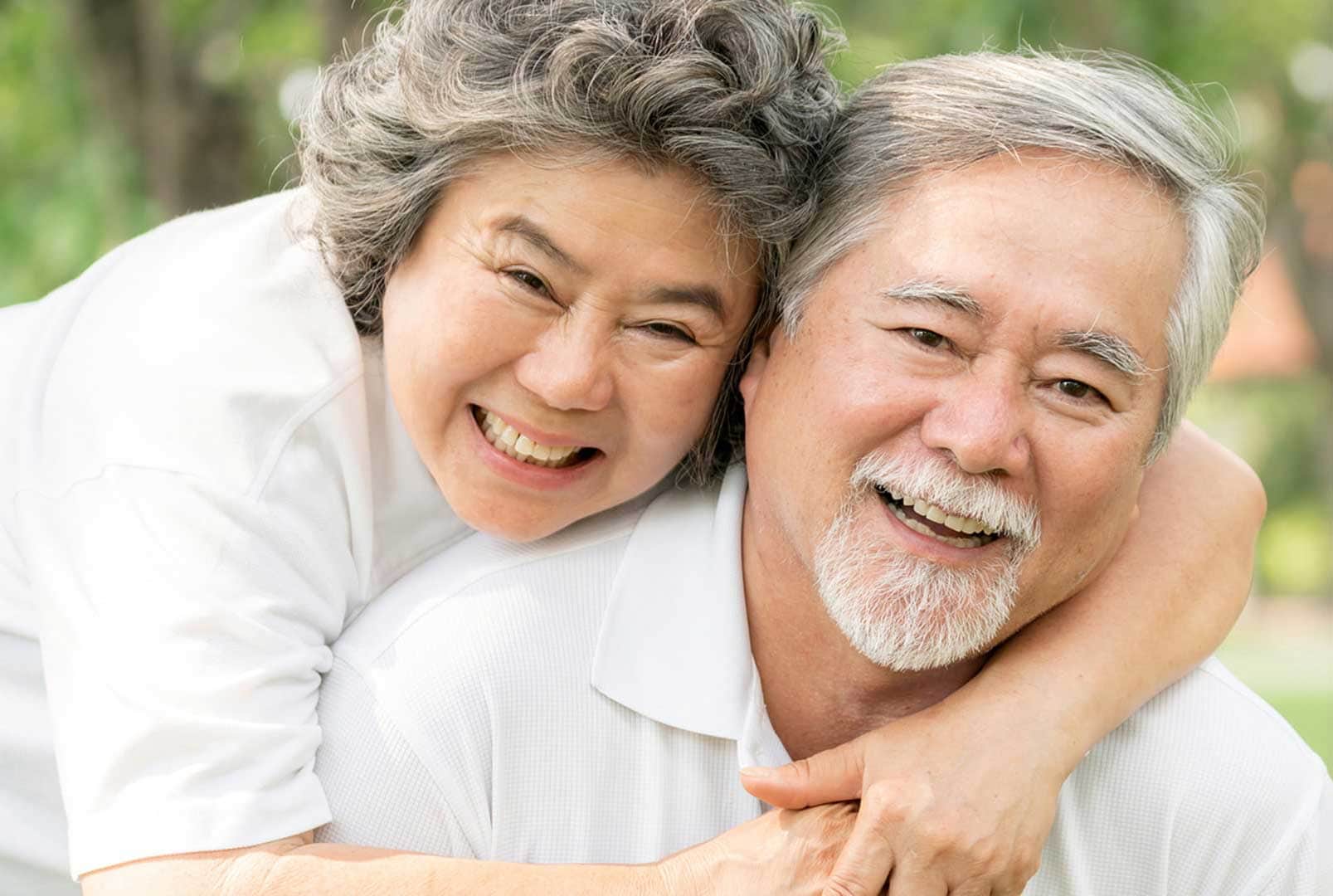 Smiling older woman behind smiling older man embracing him with her head resting on his shoulder