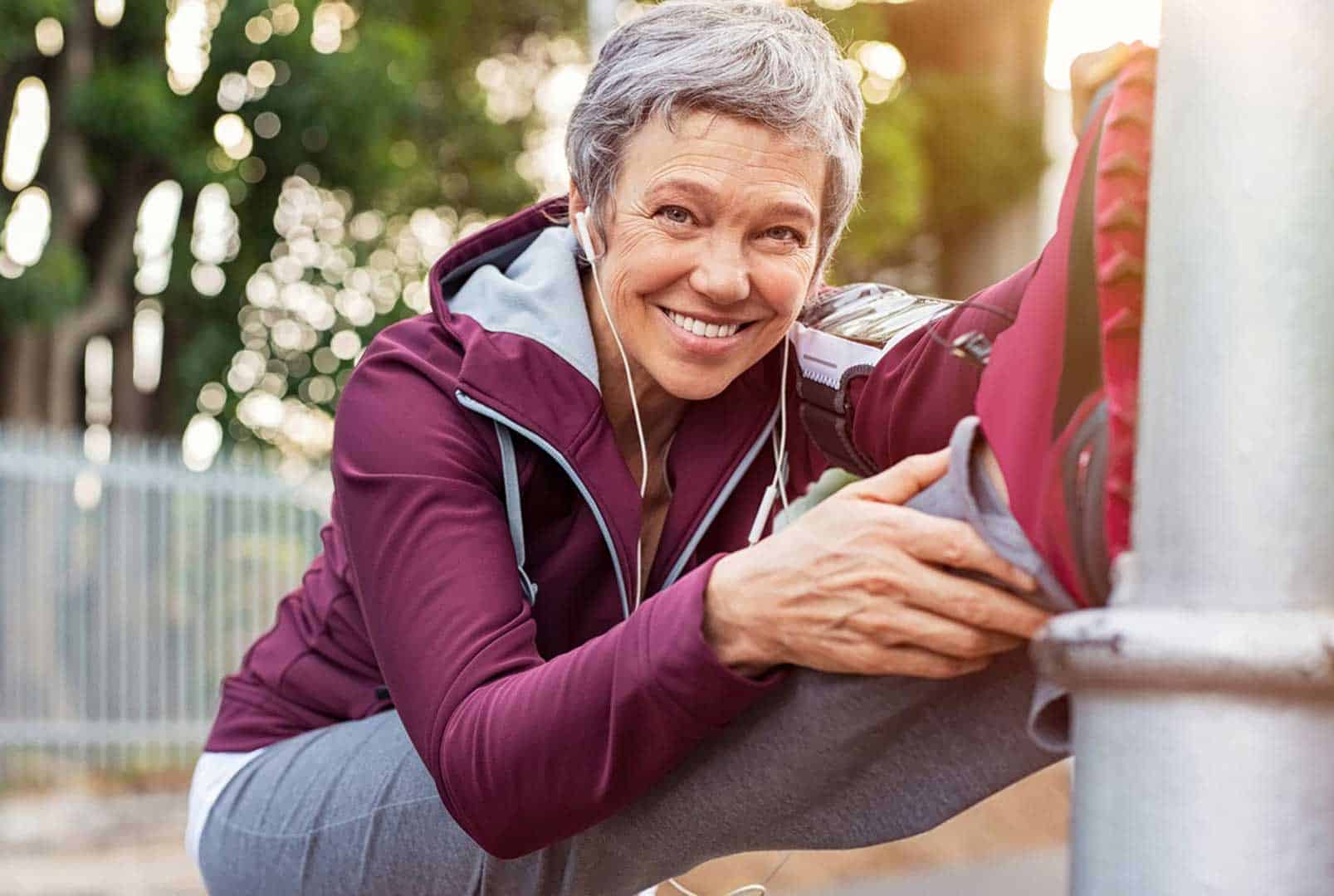 Smiling older woman wearing a tracksuit and headphones and stretching by propping a foot on a post and reaching for it