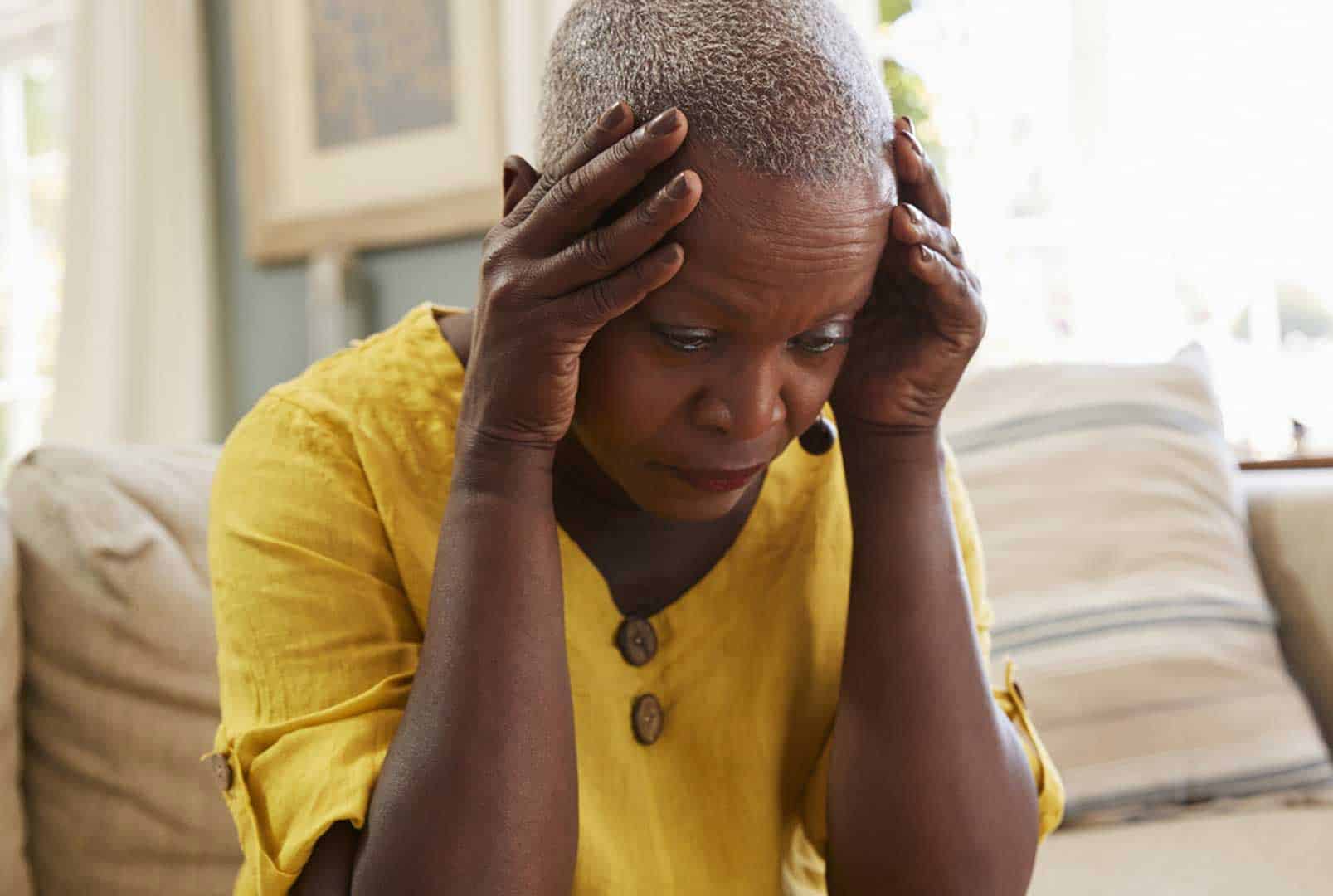 An older woman in a yellow dress sitting on a couch and holding her hands to her temples