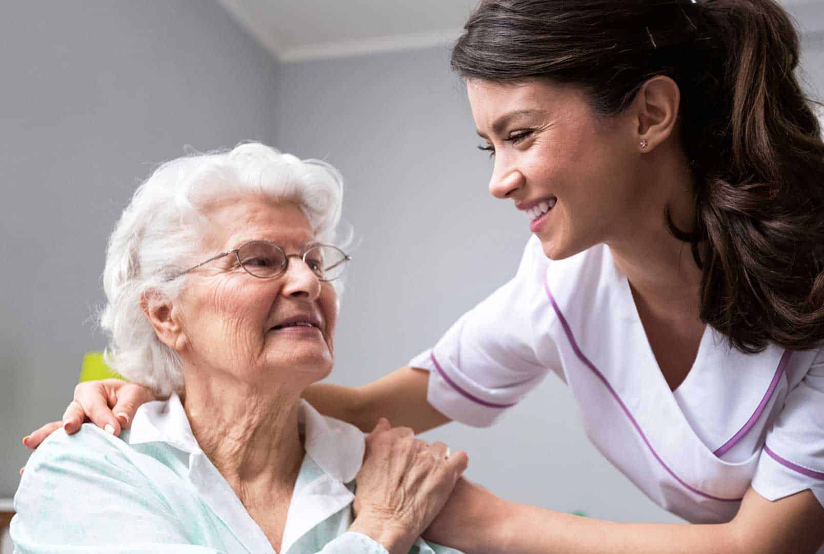 Smiling woman in a medical uniform leaning forward to embrace an older woman who is sitting in a wheelchair